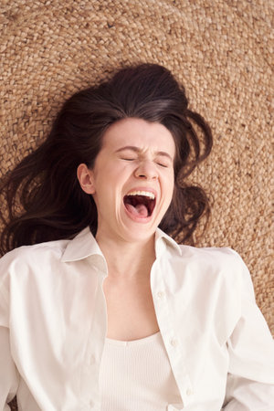 Expressive woman in white shirt laughing joyfully on textured brown woven fabric background.の写真素材