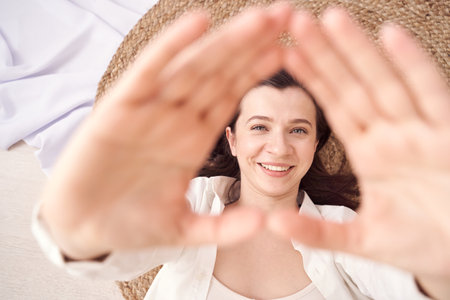 Woman lying down smiling with hands framing face in natural light setting on a cozy woven mat.の写真素材