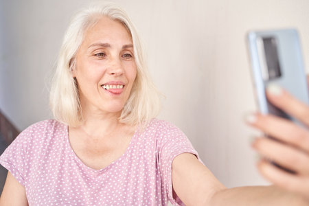 Elderly woman with gray hair taking a selfie with smartphone while smiling in relaxed casual outfit...