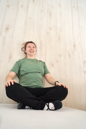 Woman smiling in green t-shirt practicing yoga seated on floor with a light wooden background.の写真素材