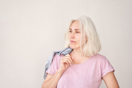 Confident mature woman with gray hair in casual outfit holding denim jacket on shoulder against neutral background.の写真素材