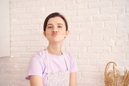 Playful woman with apron making a funny face in front of a white brick wall with wheat decoration.の写真素材