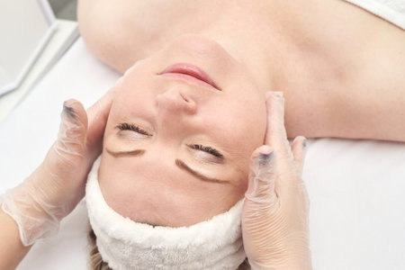 Young caucasian female receiving relaxing facial massage at spa with therapist in white gloves.の写真素材