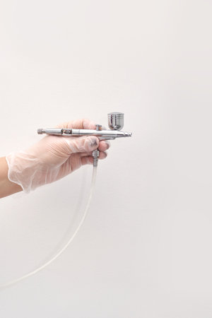 Caucasian female hand holding airbrush tool with gloves against white background.
