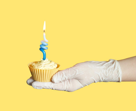 Gloved hand holding cupcake with twisted blue candle on yellow background.