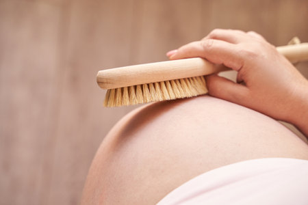 Young caucasian female using dry brush on pregnant belly for skin care and relaxation.