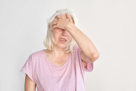 Elderly caucasian female expressing discomfort with hand on forehead against neutral background.の写真素材