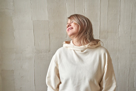 Smiling caucasian adult female with long hair in cream hoodie against textured wall background.の写真素材