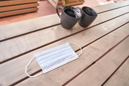 Disposable face mask on wooden table with two black coffee mugs.
