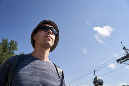 Caucasian male adult with sunglasses and hat enjoying a sunny day near cable cars.