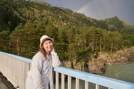 Smiling young caucasian woman on scenic bridge with rainbow and forested mountain landscape.の写真素材
