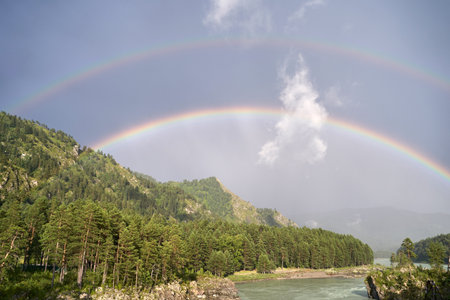 Double rainbow over lush forested mountains and river under cloudy sky.の写真素材