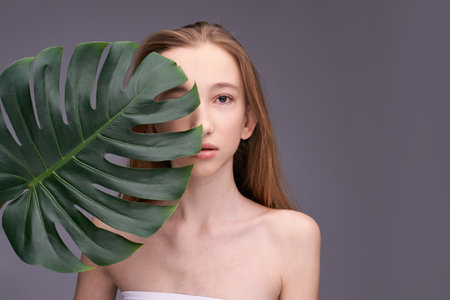 Young caucasian female with long hair holding large green leaf against gray background.の写真素材