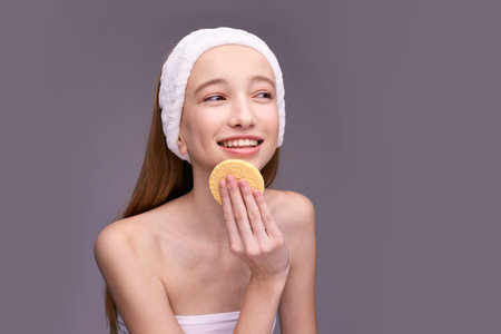 Smiling caucasian female teen using facial sponge for skin care routine with headband on grey background.の写真素材