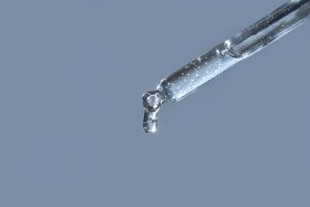 Close-up of transparent liquid drop from glass pipette against light blue background.の写真素材