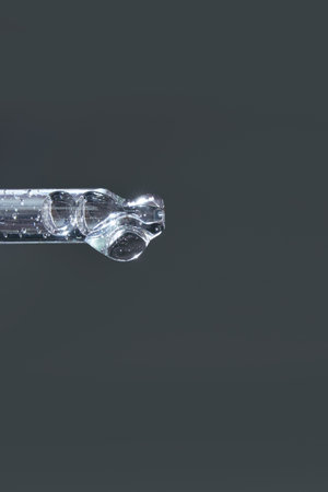 Close-up of transparent glass pipette against plain gray background with liquid droplets inside.の写真素材