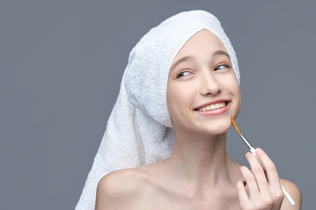 Young caucasian female applying face mask with brush smiling in white towel on gray background.の写真素材
