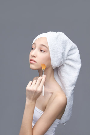Young caucasian female applying makeup with brush in towel turban against gray background.の写真素材