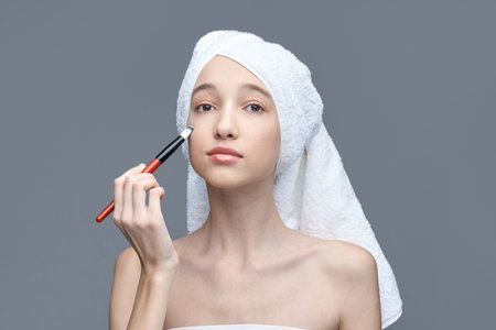Young caucasian female applying makeup with brush and towel wrapped around head.の写真素材