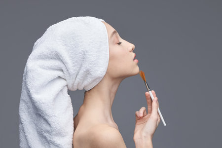 Young caucasian female applying makeup with brush in towel turban against gray background.の写真素材