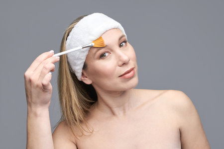 Caucasian female adult applying skincare with brush wearing headband on grey background.の写真素材