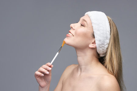Young caucasian female skincare routine with brush and headband on grey background.の写真素材