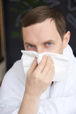 Caucasian adult male with cold symptoms using tissue for sneezing relief indoors.の写真素材