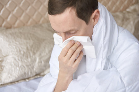 Caucasian adult male with cold or allergies blowing nose into tissue in bedroom wearing white robe.の写真素材