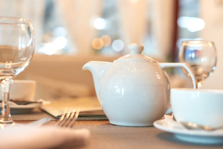 Elegant white teapot on dining table with glasses and plates in a cozy restaurant setting.の写真素材