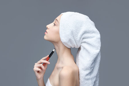 Young caucasian female applying makeup with brush on neck wrapped in towel against gray background.の写真素材