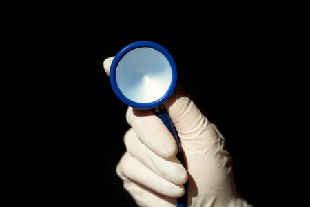 Hand wearing latex glove holding blue stethoscope against black background.の写真素材