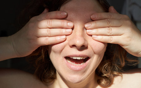 Cheerful young caucasian woman covering eyes with hands in bright sunlight.