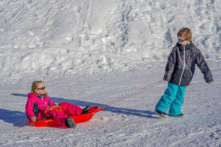 Brother pulling his sister kids toboggan sled snow. Little girl and boy enjoying sleigh ride. Child sledding. Children play outdoors in snow. Kids sled.の写真素材