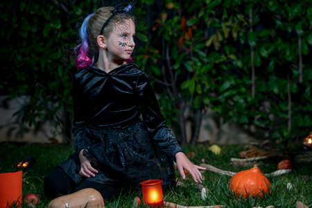 Portrait of Little girl in witch costume sitting in the night in the autumn park. Child with candy orange bucket in fall forest.の写真素材