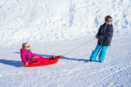 Brother pulling his sister kids toboggan sled snow. Little girl and boy enjoying sleigh ride. Child sledding. Children play outdoors in snow. Kids sled.の写真素材