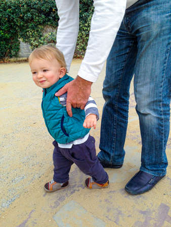 Father guiding his son with the first steps outside. Baby learning to walk, holding fathers hands. Toddler steps with the help of his father in spring park.の写真素材