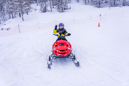 Auron, France 02.01.2021 Boy driving a red snowmobile in a winter landscape. Portrait of smiling boy in winter. Warm clothes, winter sport. Happy child on a snowmobile. High quality photoのeditorial素材