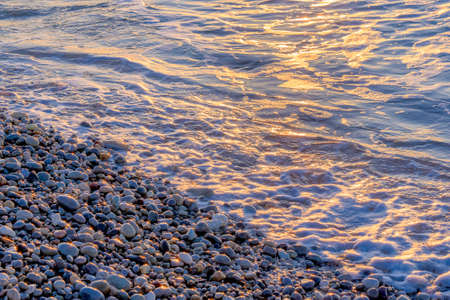 Golden pebbles on seashore in morning sun light. Sea wave on seacoast with round stones....