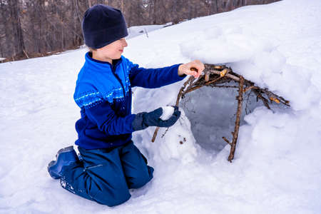 Young boy building a hut with a snowy and branches in the winter forest. Outdoor activity with...