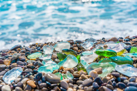 Natural polish textured sea glass and stones on the seashore. Azure clear sea water with waves. Green, blue shiny glass with multi-colored sea pebbles close-up. Beach summer background.の写真素材