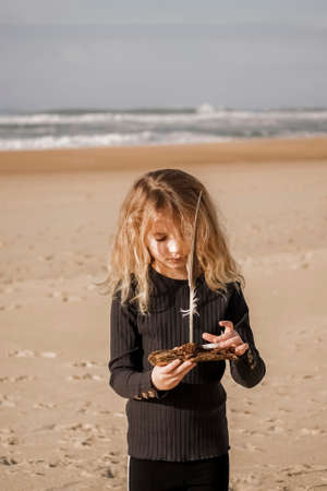 The Girl is along on the sandy shore with piece of wood and seagull feather as toys in the hands. Beach landscape on the background. High quality photoの写真素材
