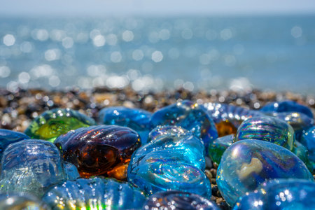 Beautiful shape glass and stones on the seashore. Azure clear sea water with waves. Green, blue shiny glass with multi-colored sea pebbles close-up. Beach summer background. High quality photoの写真素材
