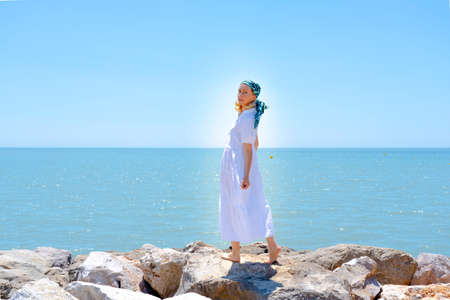 Side view of a young woman wearing a white dress and hair scarf standing on rocks on a sea