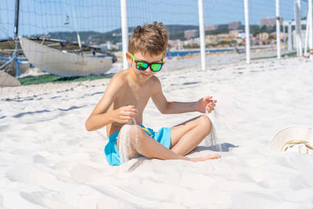 Handsome young boy in blue shorts and sunglasses sits on the white sand on the beach. Plays with white sand. High quality photoの写真素材