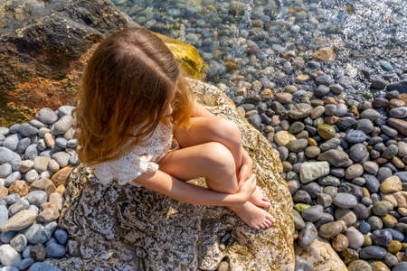 Beautiful little girl with blond hair sitting on a rock by the sea. High quality photoの写真素材