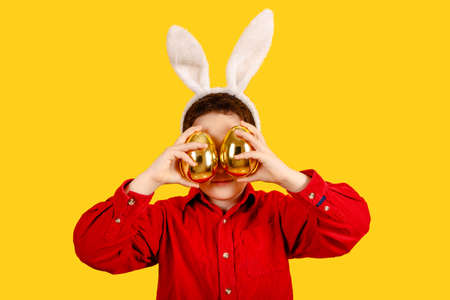 Funny child in bunny ears headband holding Golden Easter eggs on color background