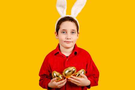 Sweet boy in bunny ears headband holding Golden Easter eggs on color background