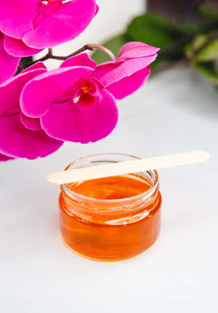 Wax or sugar paste for hair removal in a glass jar with a wooden wax spatula in molten with orchids flowers on a white background. Copy space. Depilation and beauty conceptの写真素材
