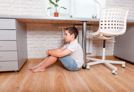 Offended upset boy teenager sitting on the floor. Schoolboy torturing of bullying. Teen boy sitting under the desk in the nursery. Teenager one cries. Problem of the relations of parents and childrenの写真素材
