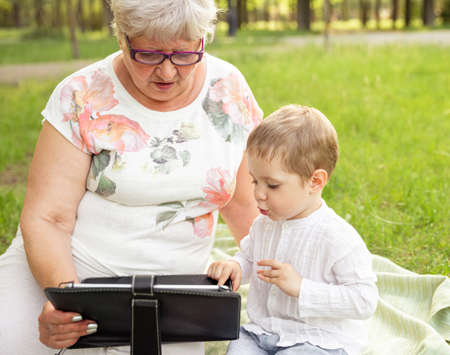 Little boy teach grandma in glasses to use modern smart devices outdoors. Communication of young and senior generation. Enjoy leisure time with family. Advanced older generation and technologyの写真素材